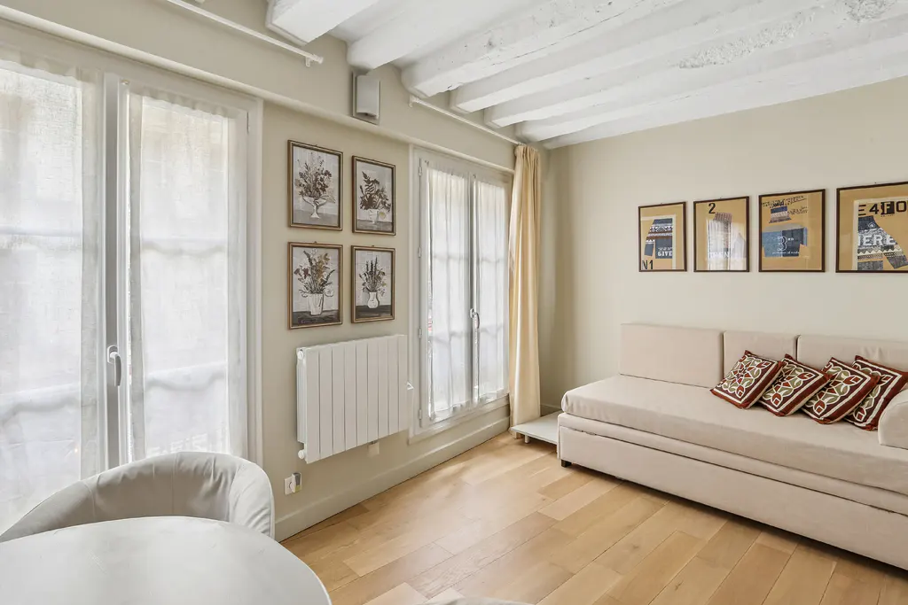 Bright living room with wood floors, white sofa with patterned pillows, and framed art on the walls. Windows with sheer curtains let in natural light.