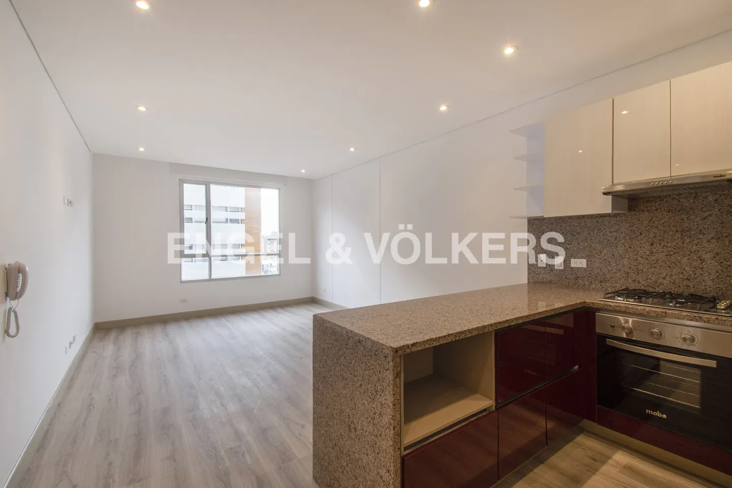 A modern apartment interior with white walls, light wood floors, and a granite kitchen island with red cabinets. A window provides natural light.