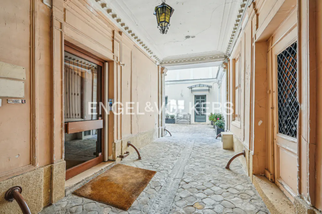 Arched walkway with stone floor and peach walls leads to a courtyard with a bench and plants. An ornate lantern hangs from the ceiling.