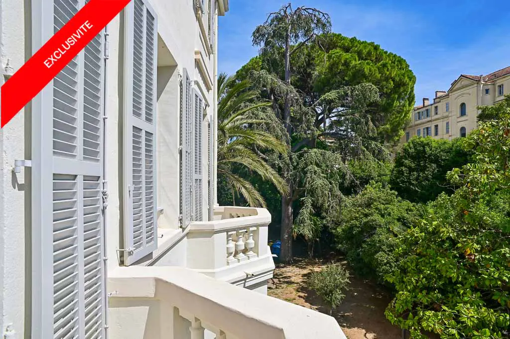 Balcony view of a white building with gray shutters, a red "Exclusivite" banner, and lush green trees in the background.