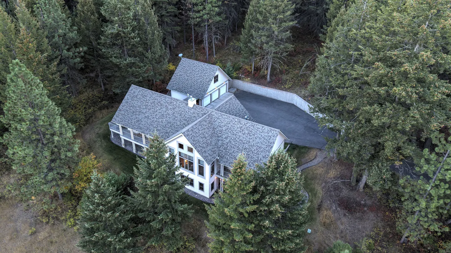 Aerial view of a large, light gray house with a gray roof, surrounded by tall green trees. A driveway leads to a garage.