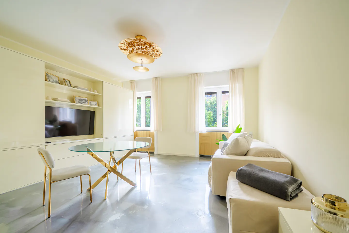 Bright living room with a glass table, gold chairs, a beige sofa, and a built-in TV unit. Two windows with sheer curtains let in natural light.