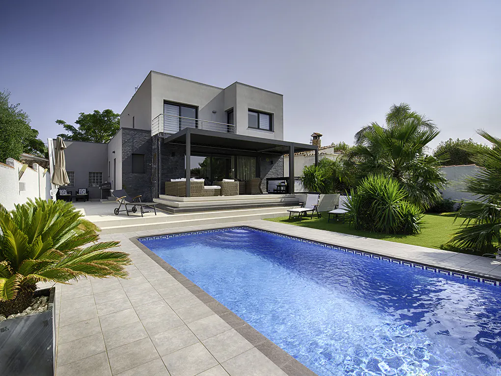 Modern two-story house with a blue swimming pool, patio furniture, and green lawn under a clear blue sky.