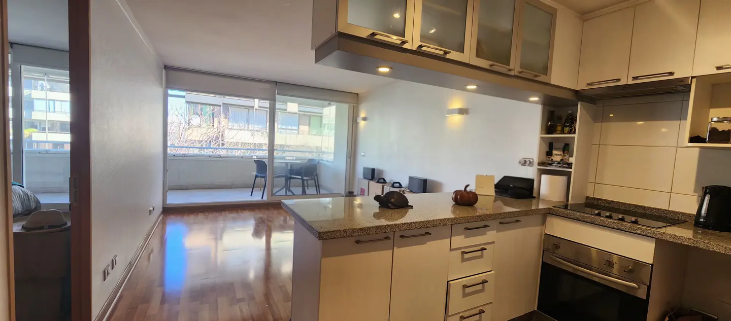 A bright kitchen with white cabinets, granite countertops, and stainless steel appliances. A balcony with a table and chairs is visible through sliding glass doors.