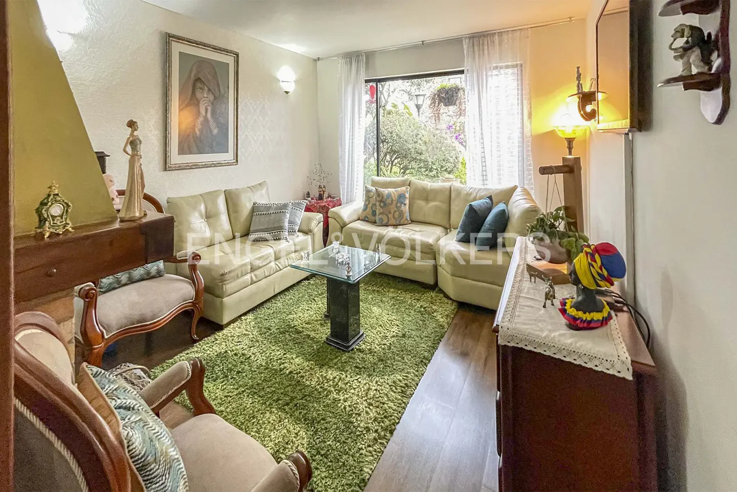 A living room with cream sofas, a glass table on a green rug, and a view of a garden through a window.