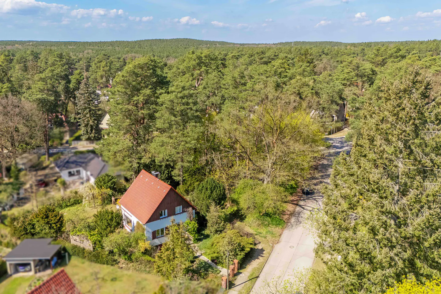 Aerial view of a two-story house with a red roof surrounded by lush green trees and a road.