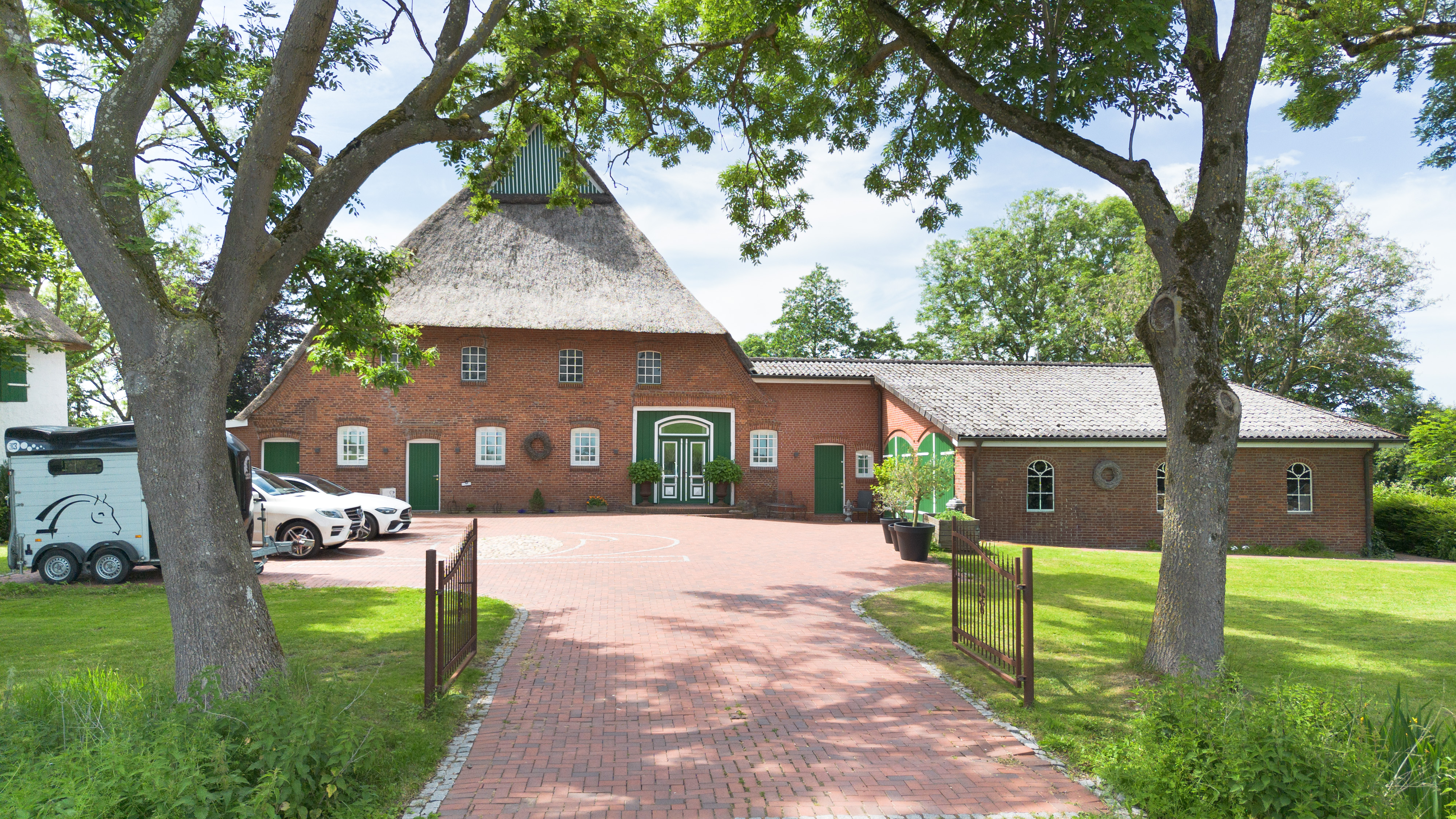 A large red brick farmhouse with a thatched roof, a paved courtyard, and a horse trailer parked under mature trees. [AI-generated text]