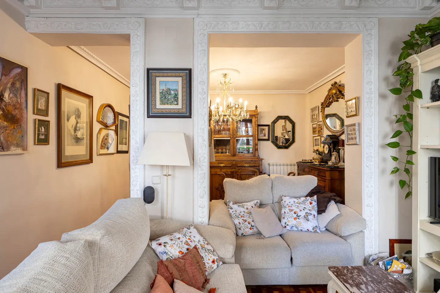 Living room with beige sofas, floral pillows, and ornate white trim. A chandelier hangs over a wooden cabinet in the background. Walls are decorated with framed art.
