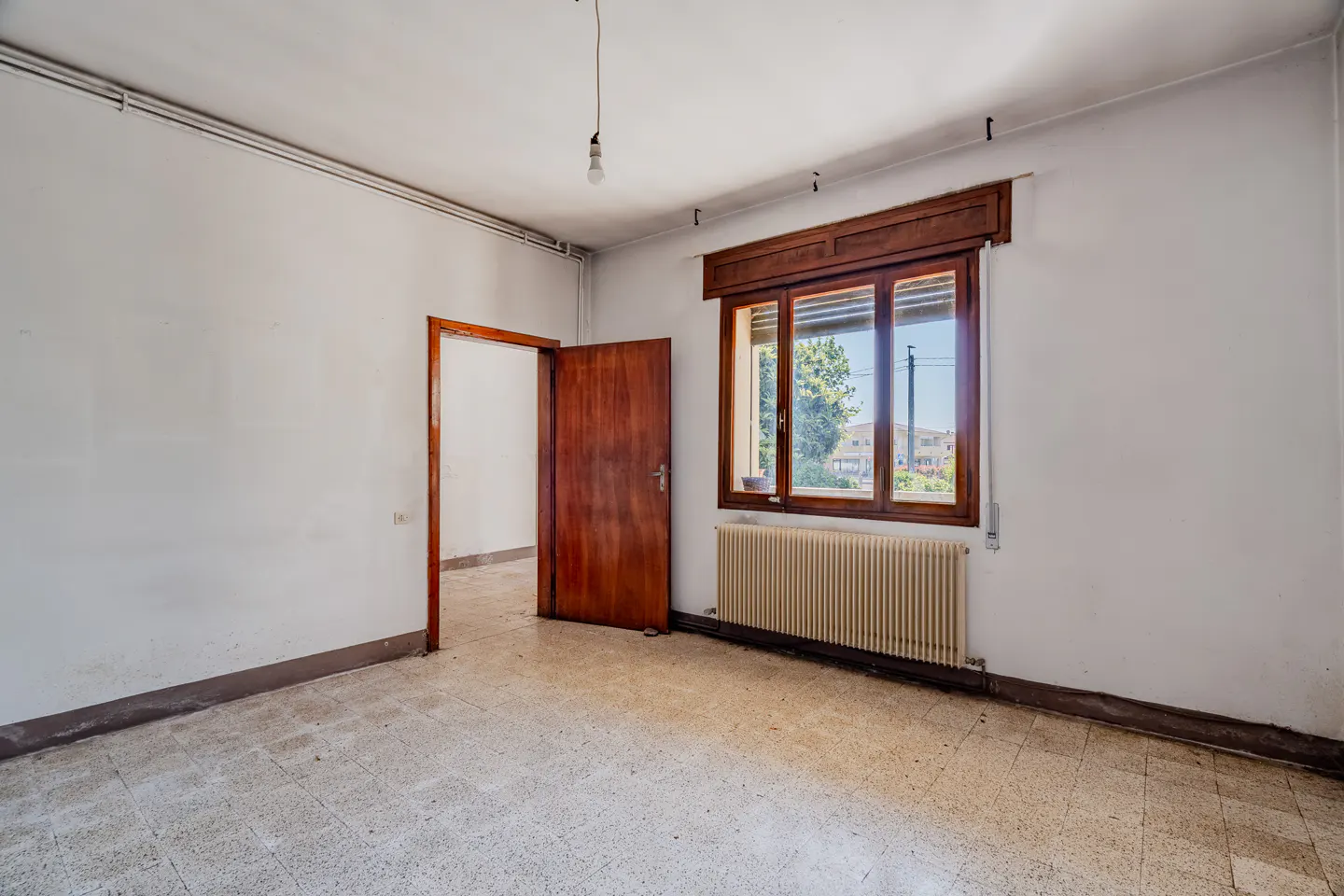 A vacant room with white walls, a brown window frame, a radiator, and a brown door.