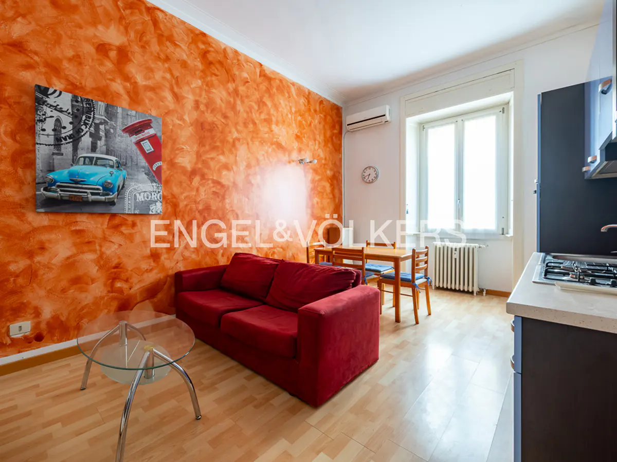 Living room with orange textured wall, red sofa, glass table, and dining set. A blue car art piece hangs on the wall.