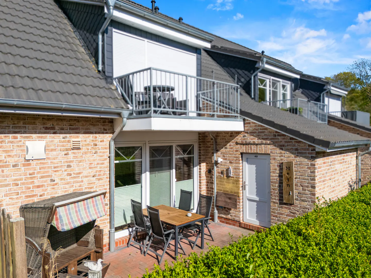 Brick townhouse with a patio featuring a table, chairs, and a wicker beach chair. A balcony with seating is above the patio.