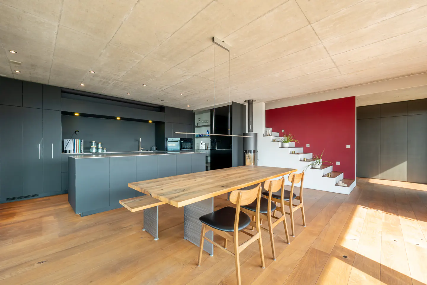 Modern kitchen with dark grey cabinets, a long wooden dining table, and a staircase against a bold red accent wall.