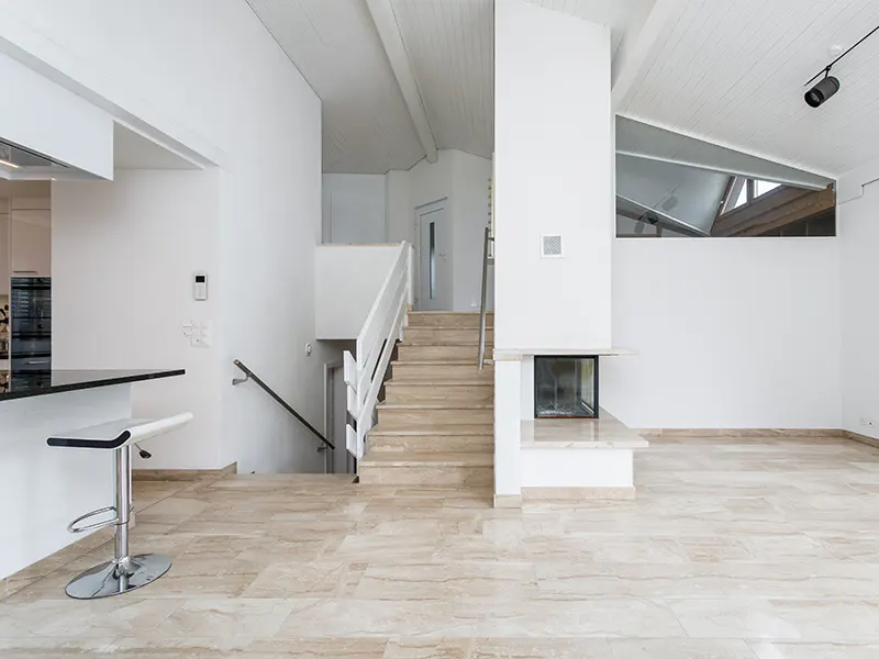 Bright, modern interior with beige tile floors, white walls, and a staircase. A fireplace and a kitchen bar with a stool are visible.