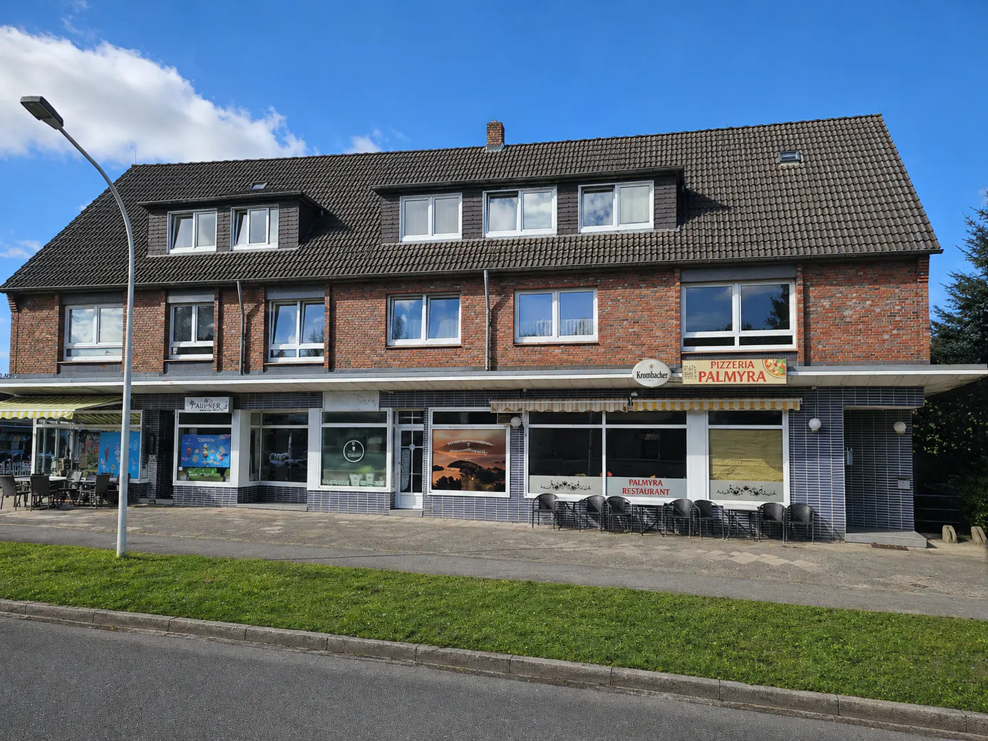 Two-story brick building with a dark roof, housing businesses including Pizzeria Palmyra, under a blue sky.