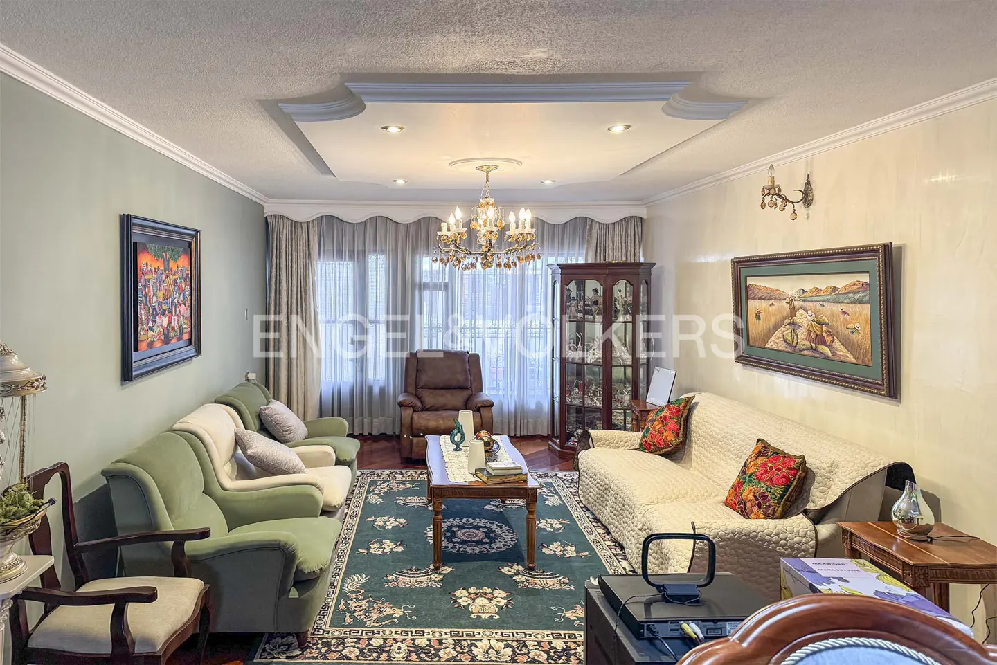 A living room with green and beige sofas, a brown recliner, and a patterned rug. A chandelier hangs from the ceiling.