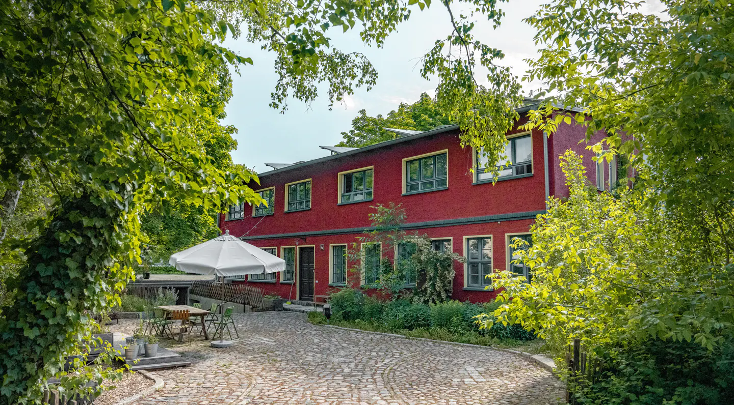 Red two-story building with green trim, surrounded by trees. A white umbrella covers a table and chairs on a stone patio.