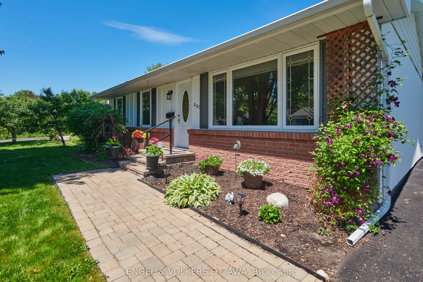 Exterior of a white bungalow with a brick base, stone walkway, and a garden bed featuring potted flowers and a trellis.
