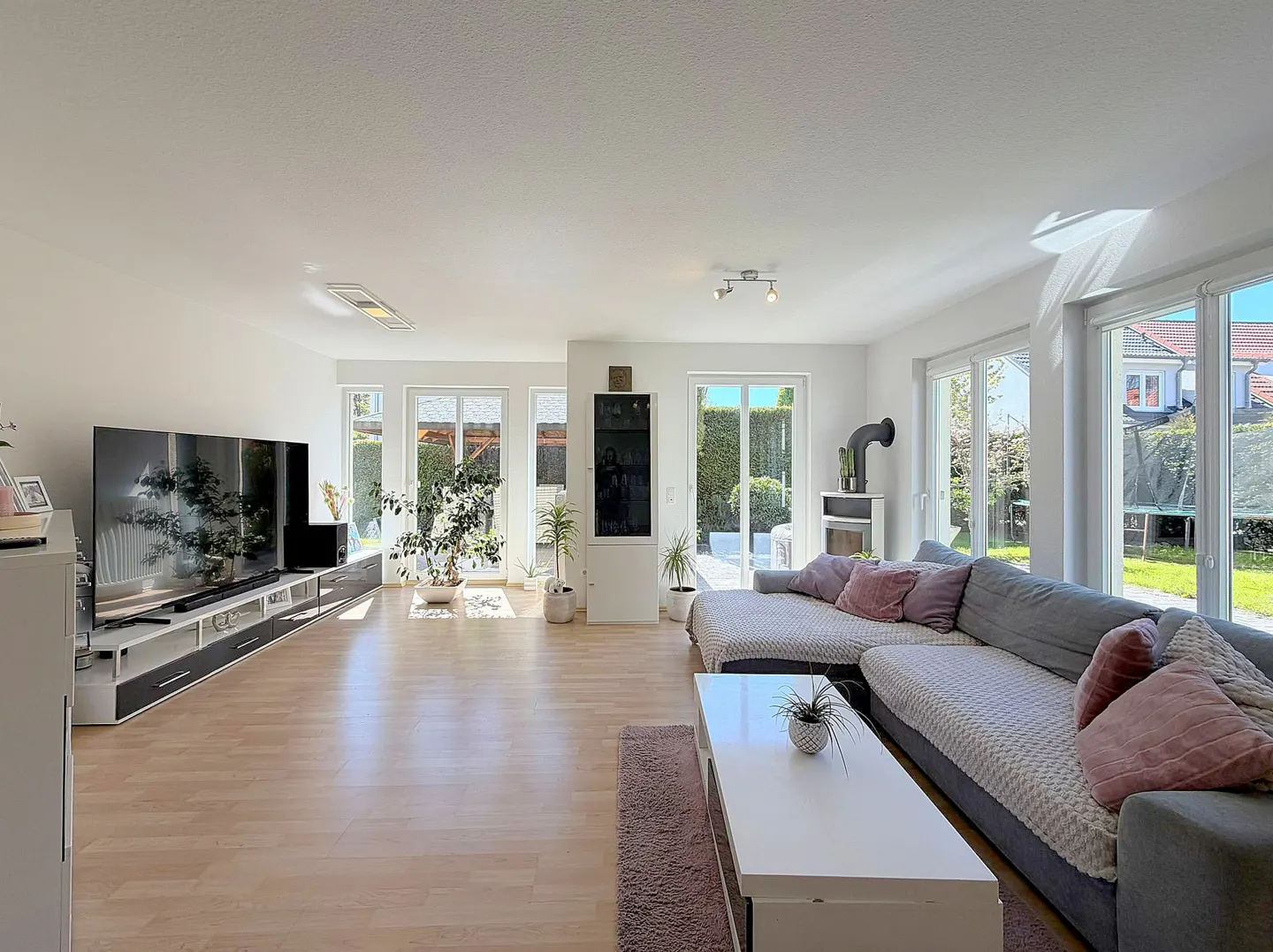 Bright living room with wood floors, a gray sectional sofa with pink pillows, and a large TV. Sunlight streams through the glass doors to the backyard.