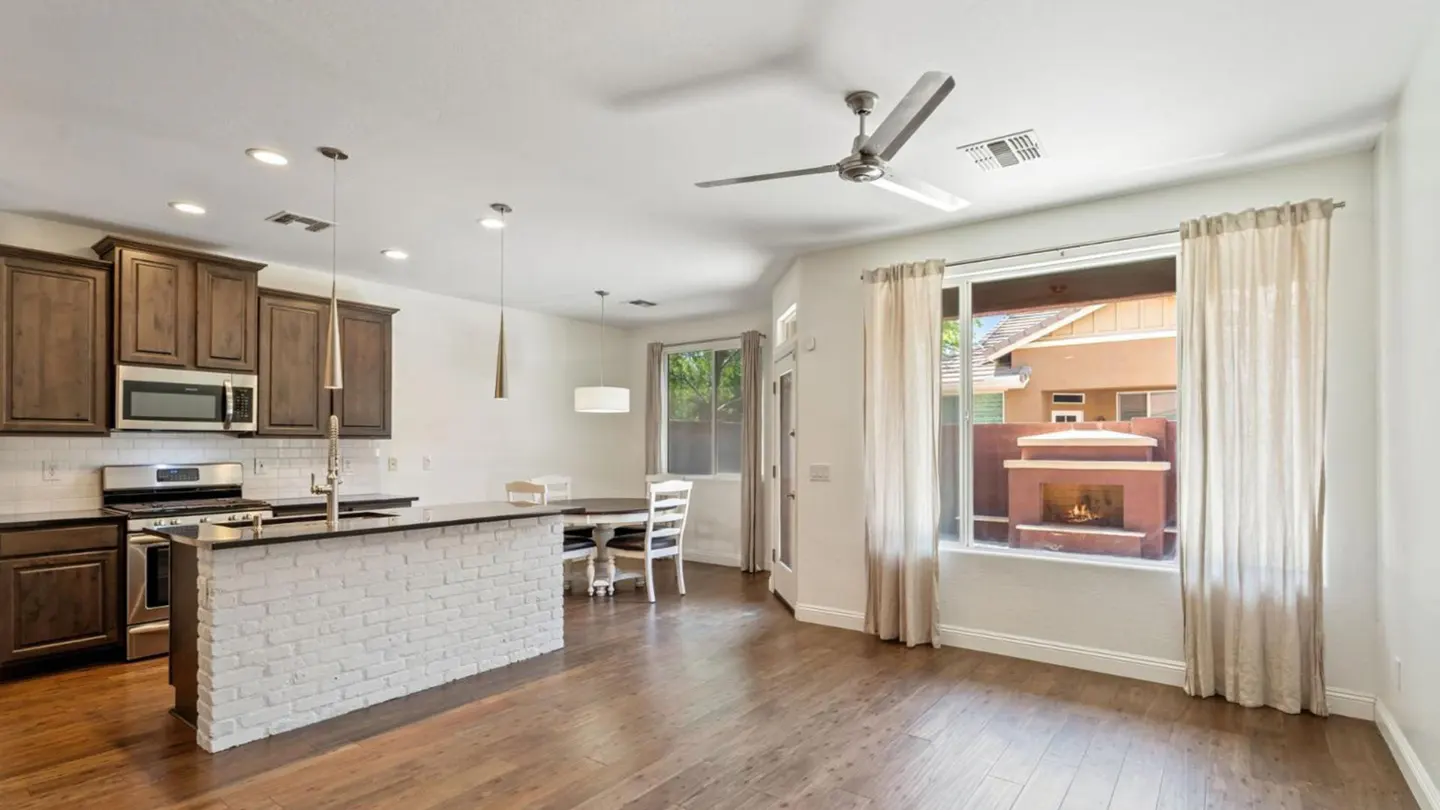 A bright, modern kitchen with dark wood cabinets, white brick island, and hardwood floors. A dining area and window with curtains are visible.