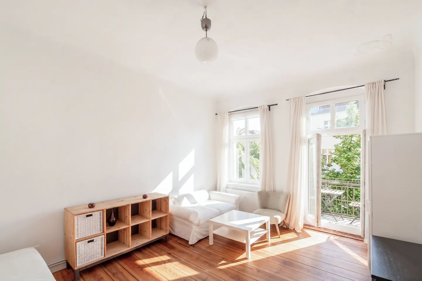 Bright living room with hardwood floors, white walls, and natural light. Features a sofa, chair, table, shelf, and balcony.