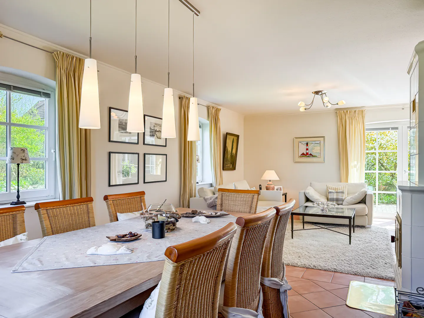 Bright, open-concept living and dining area. A long wooden table with wicker chairs sits under pendant lights. White sofas and a rug are in the background.