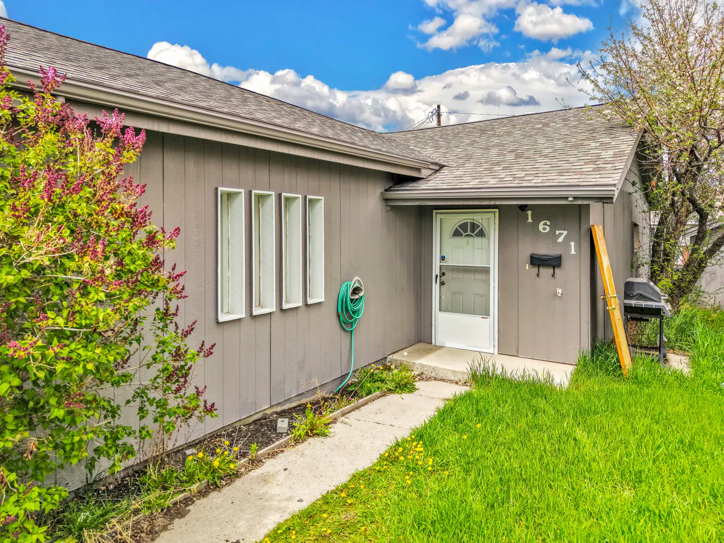 A gray house with a white door and the number 1671 above the mailbox. A green hose is coiled on the side of the house.