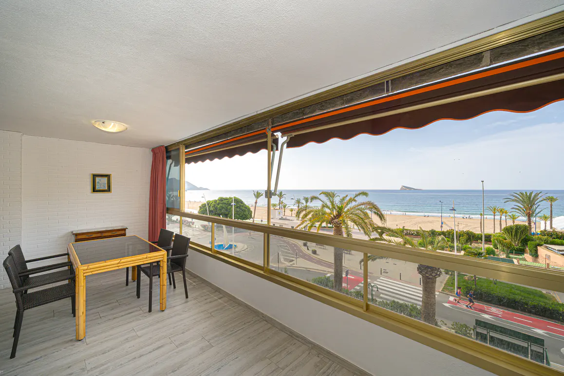 Balcony view of a beach with palm trees. Table and chairs on the balcony. Brown awning above the window.