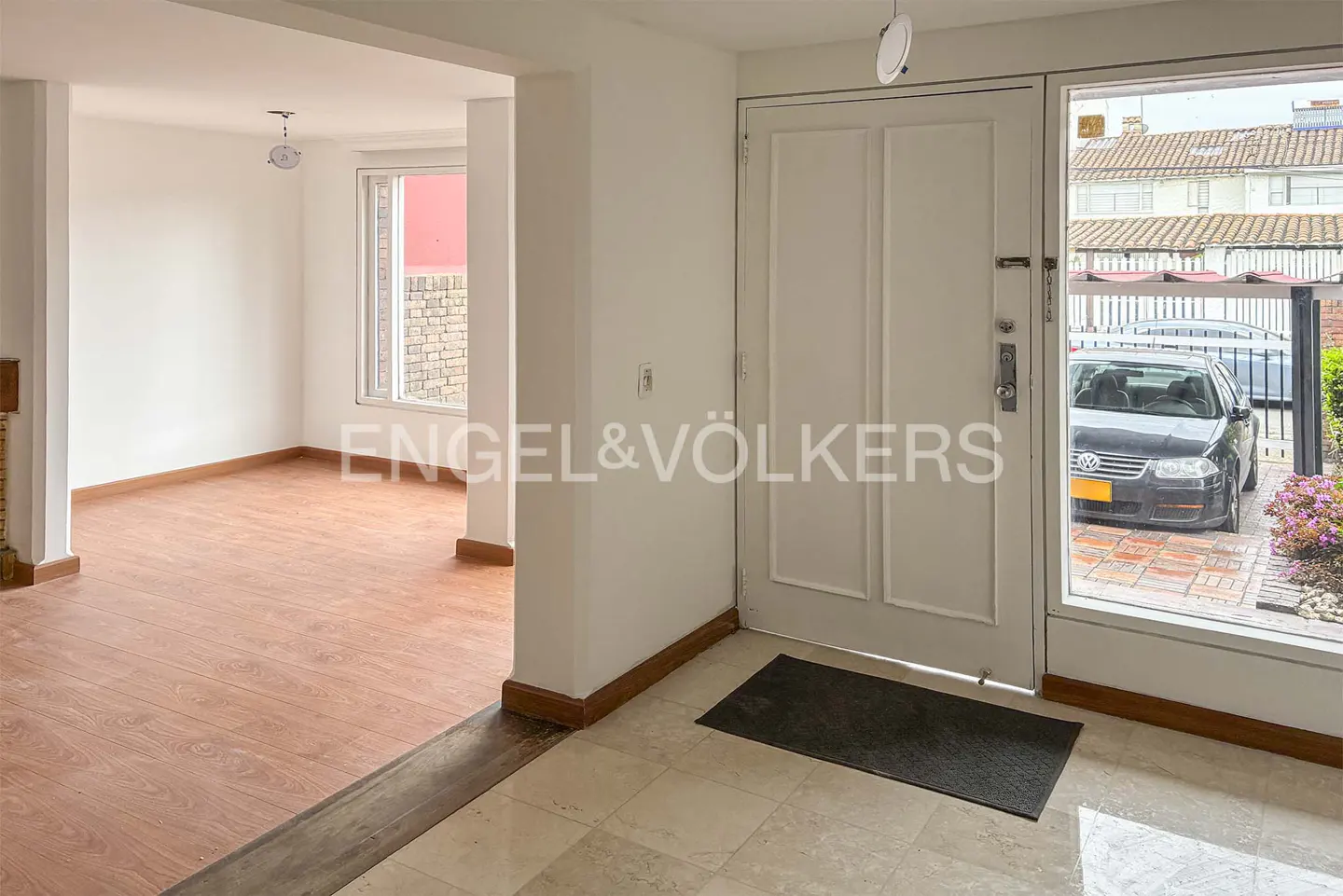 A bright foyer with a white front door, a black car visible through the glass, and an open living room to the left.