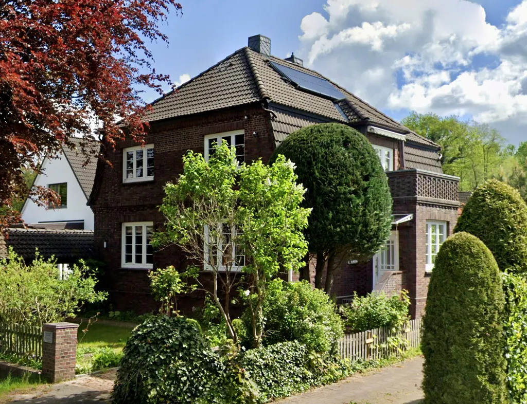 A two-story brick house with a brown tiled roof and solar panels, surrounded by green trees and bushes.