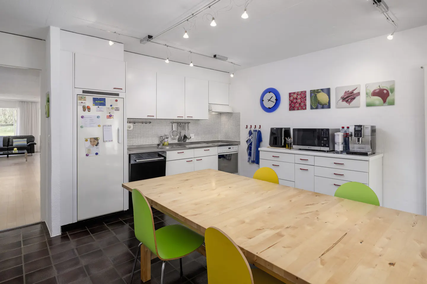 Bright kitchen with white cabinets, appliances, and a large wooden table with green and yellow chairs. Fruit-themed art hangs on the wall.