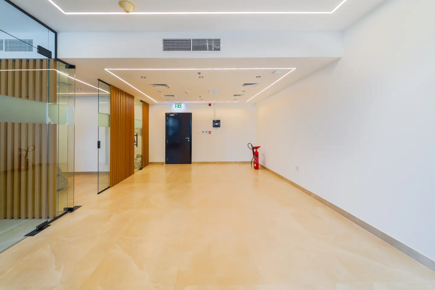 Bright, empty office space with glass-walled rooms, a black door with exit sign, and a red fire extinguisher against a white wall.