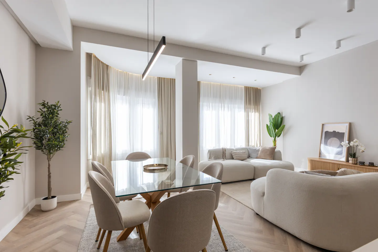 Bright, modern living and dining area with beige sofas, chairs, and curtains. A glass table sits on a herringbone wood floor.