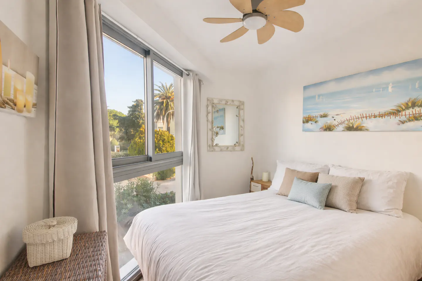 Bright bedroom with a white bed, pillows, and a beach painting. A large window shows trees and a palm tree. A woven basket sits on a wicker chest.