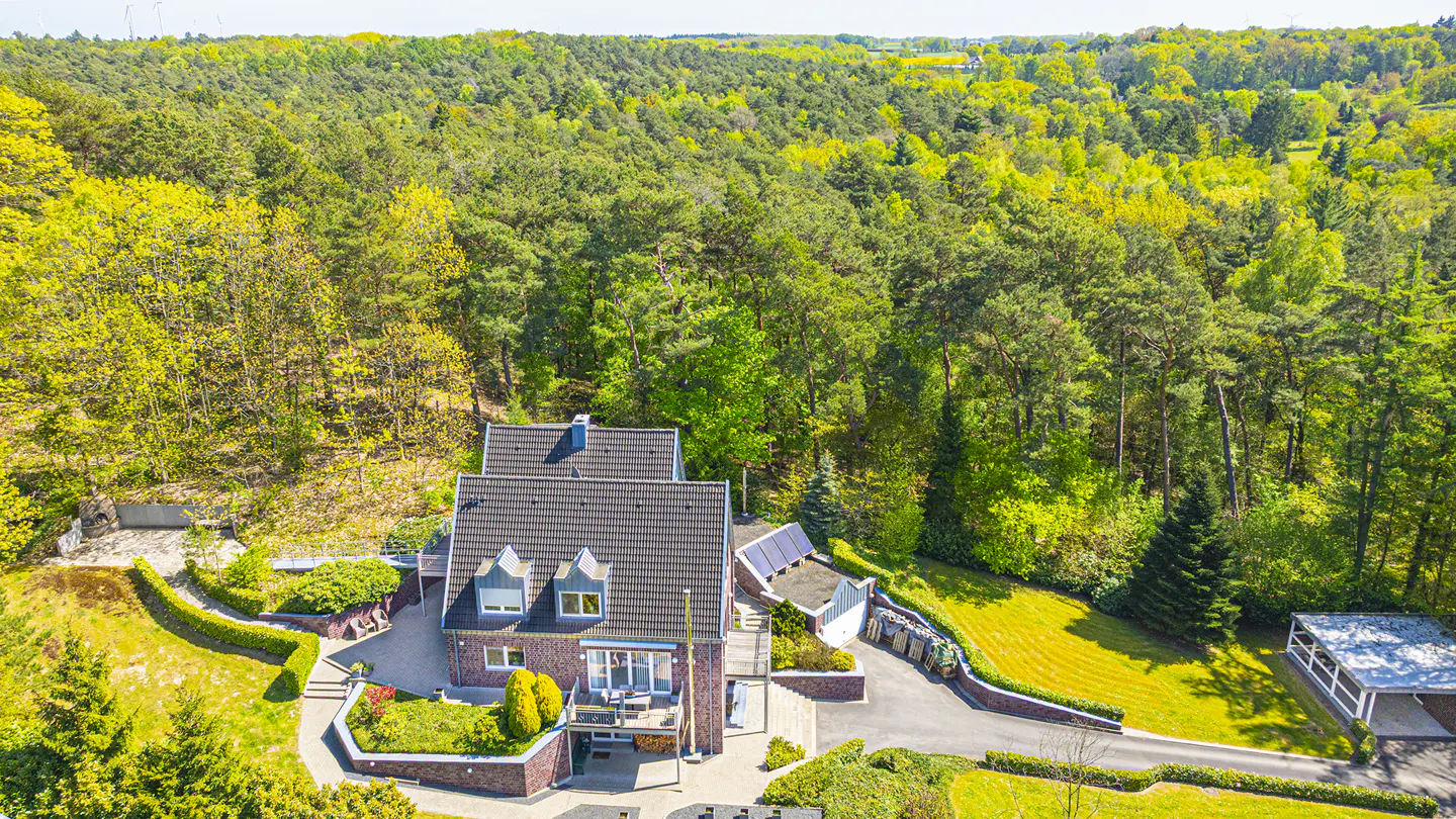 Aerial view of a brick house with a gray roof, surrounded by lush green trees and manicured lawns on a sunny day.