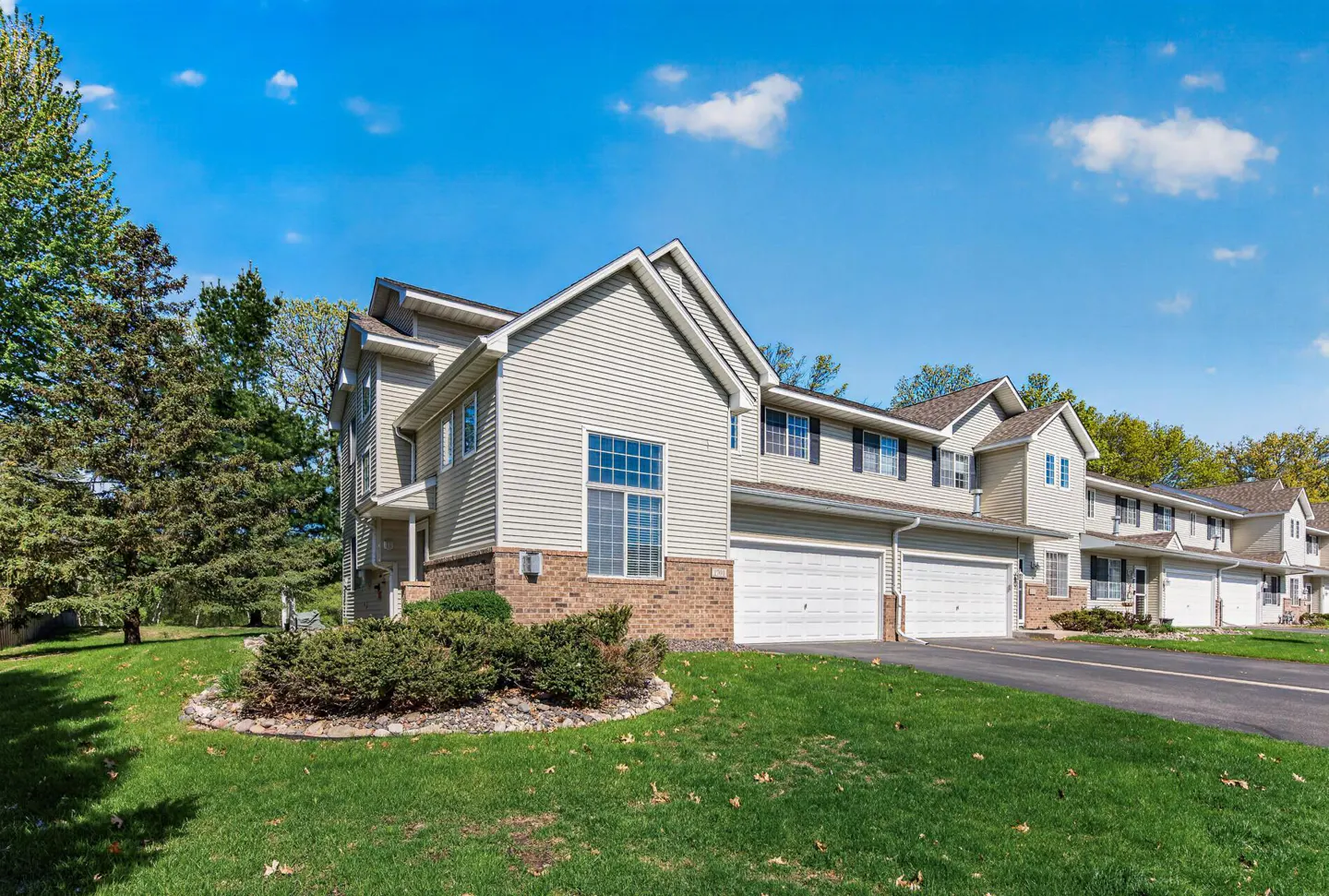 Beige two-story townhouses with white garage doors, green lawns, and trees under a blue sky.