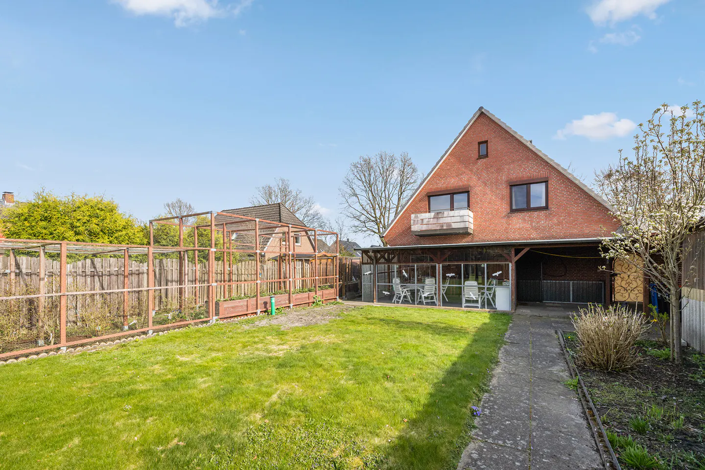 A red brick house with a balcony overlooks a green lawn and garden with a wooden fence and pergola.
