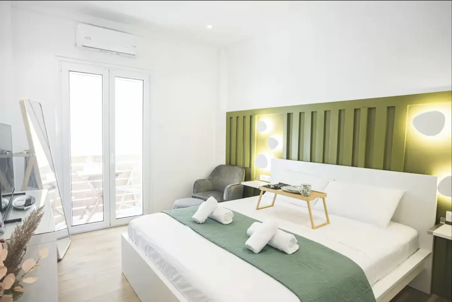 Bright bedroom with white walls, a bed with a green runner, and a green headboard with lights. A balcony is visible through glass doors.
