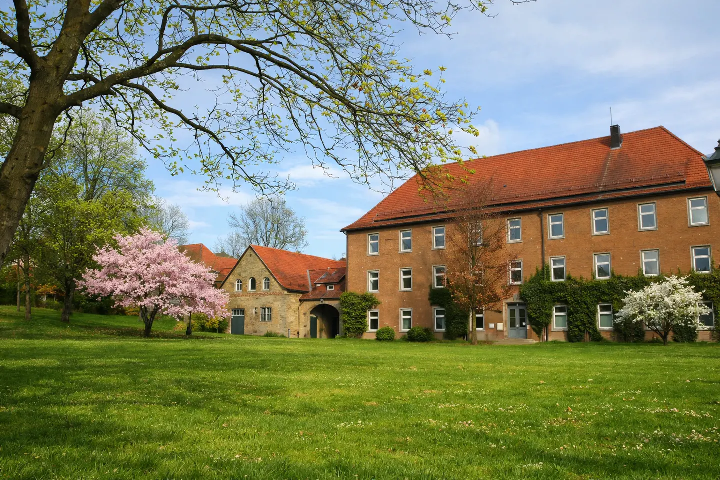 A large, tan building with a red tile roof sits on a green lawn with trees and flowering bushes.