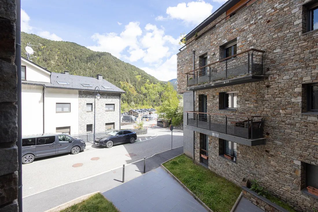 View of a stone building with balconies, a street with parked cars, and green mountains in the background.