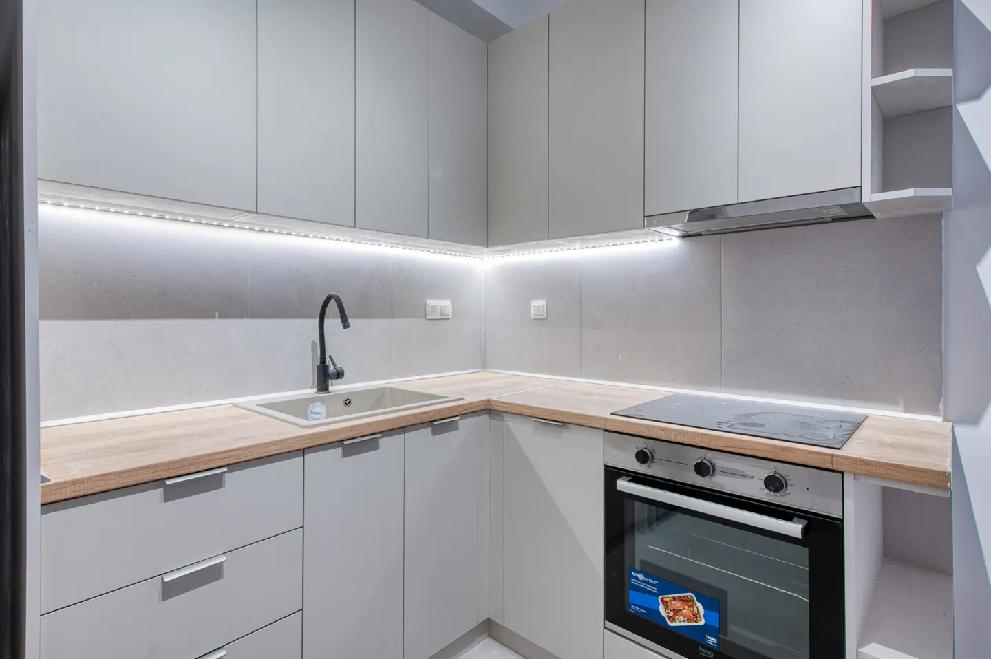 Modern kitchen with gray cabinets, wood countertops, and stainless steel oven. Black faucet over a beige sink. LED strip lighting under cabinets.