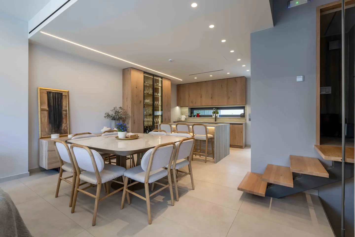 Open-concept dining and kitchen area with light wood cabinets, island seating, and a marble-topped dining table with white chairs.