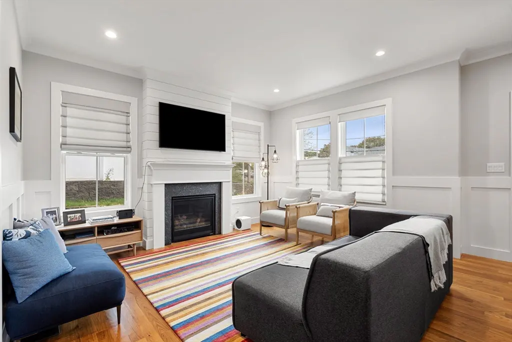 Bright living room with gray sofa, blue chaise, and two chairs. A fireplace with a TV above it and a colorful striped rug add warmth.