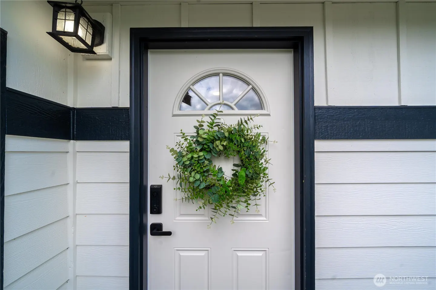 White front door with a green eucalyptus wreath, black trim, and a modern outdoor light on white horizontal siding.