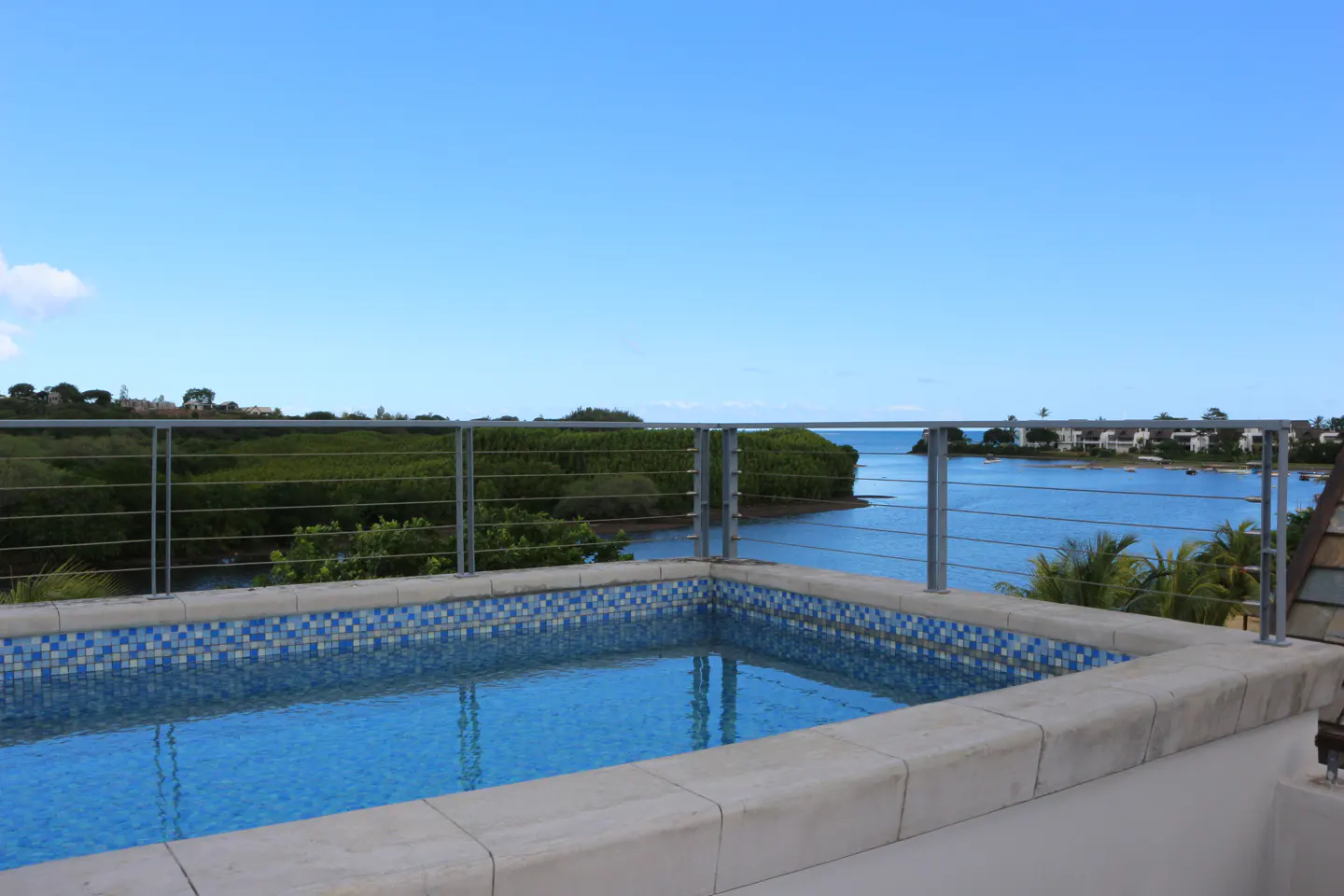 Rooftop pool with blue mosaic tiles, overlooks a bay with green trees and blue water under a clear blue sky.