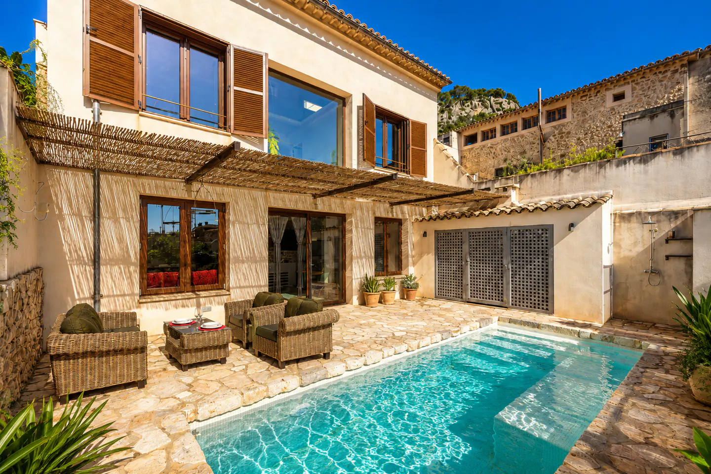 A sun-drenched courtyard with a turquoise pool, stone patio, and wicker furniture. The building has brown shutters and a bamboo awning.