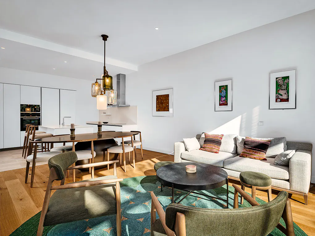 Bright, modern living space with white walls, wood floors, and a green rug. A gray sofa, dining table, and kitchen are visible.