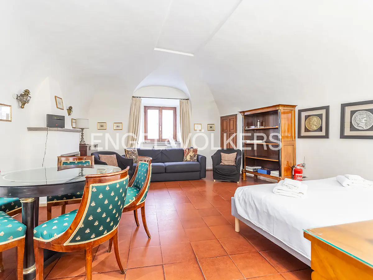 Open-concept living space with a bed, sofa, table, and chairs on a terracotta tile floor. The walls are white and the ceiling is arched.