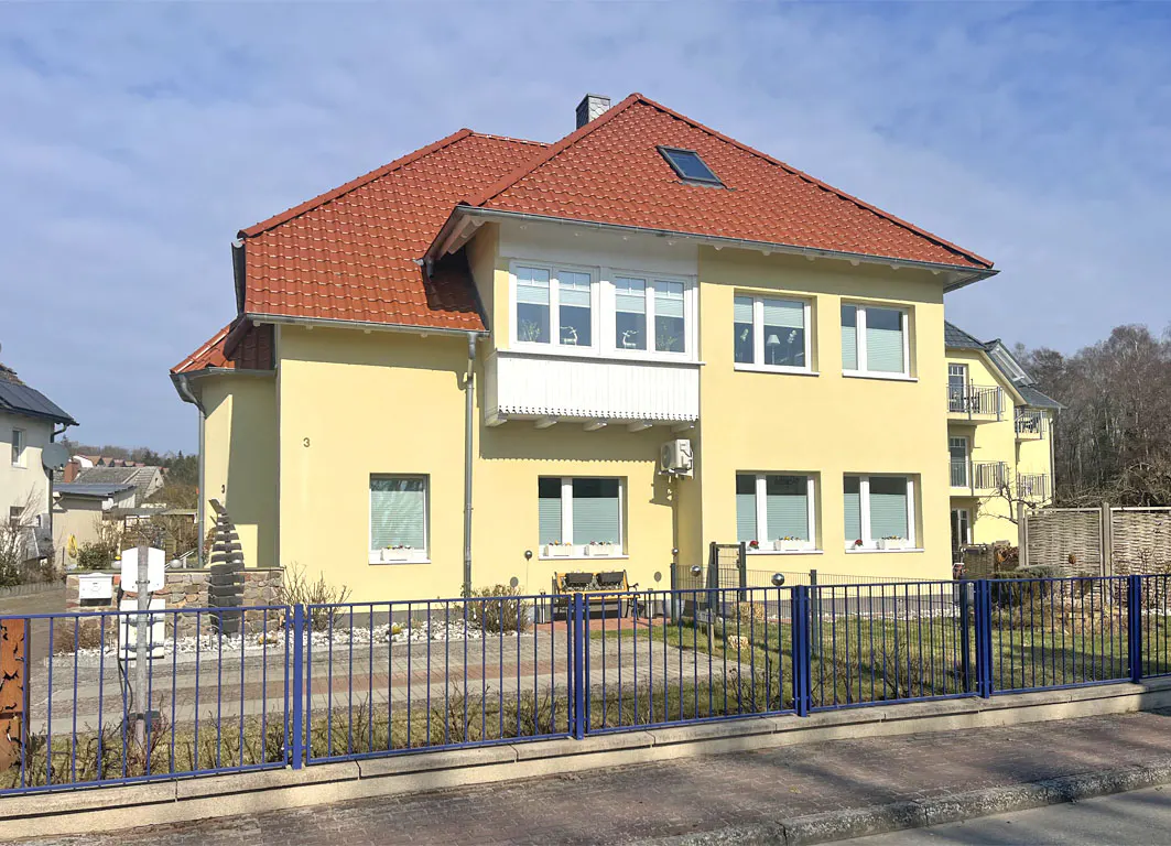 Two-story yellow house with a red tile roof and blue metal fence in front.