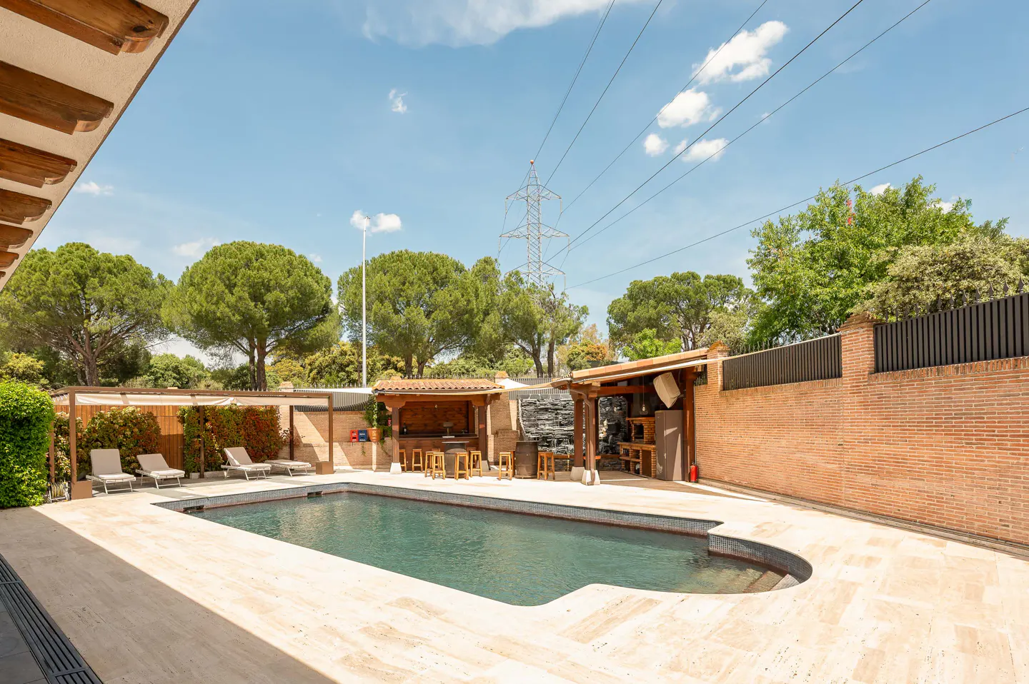 Outdoor pool area with lounge chairs, bar, and waterfall feature. Brick wall and green trees surround the space under a blue sky.