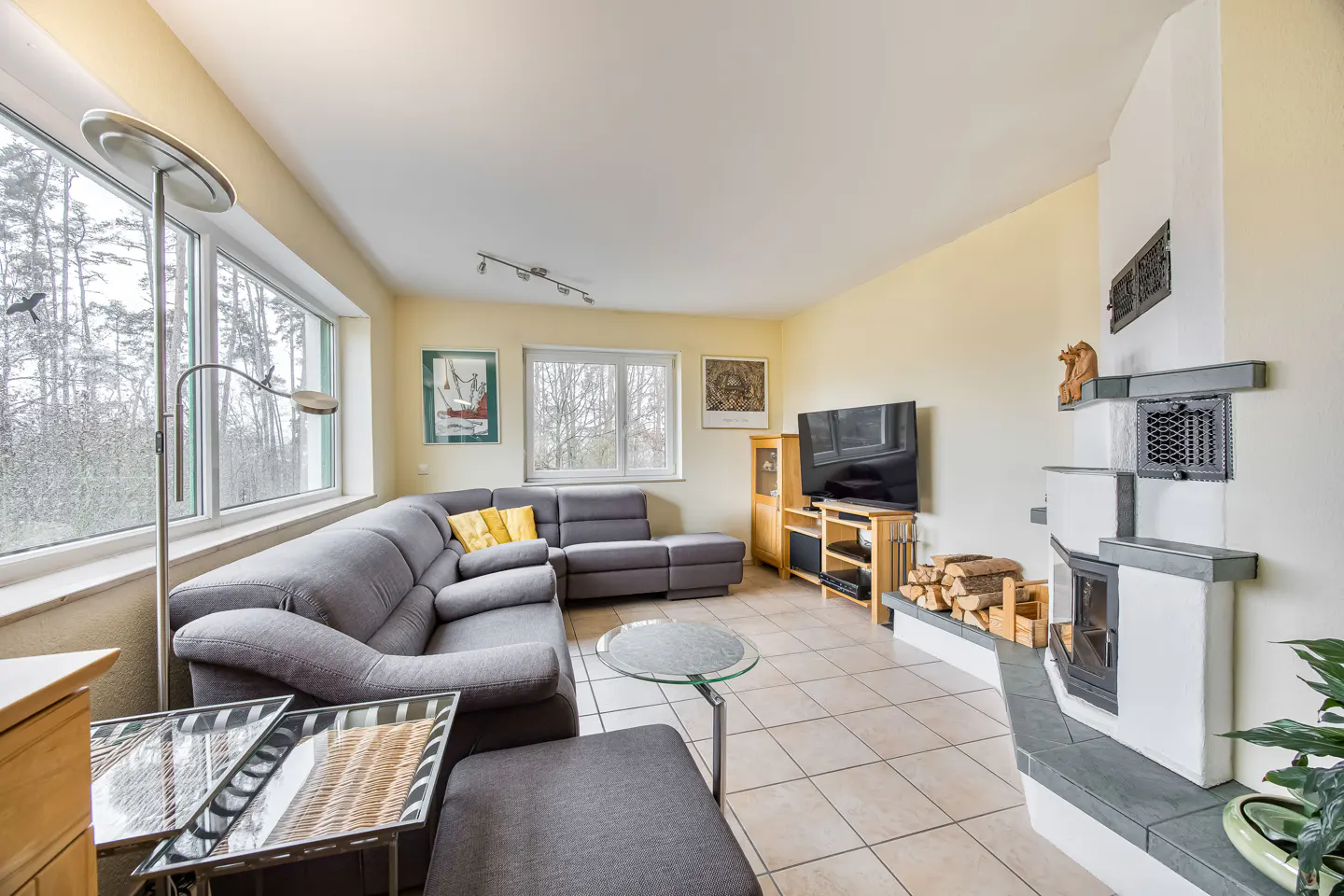 Living room with gray sectional sofa, yellow pillows, fireplace, TV, and large windows overlooking trees.